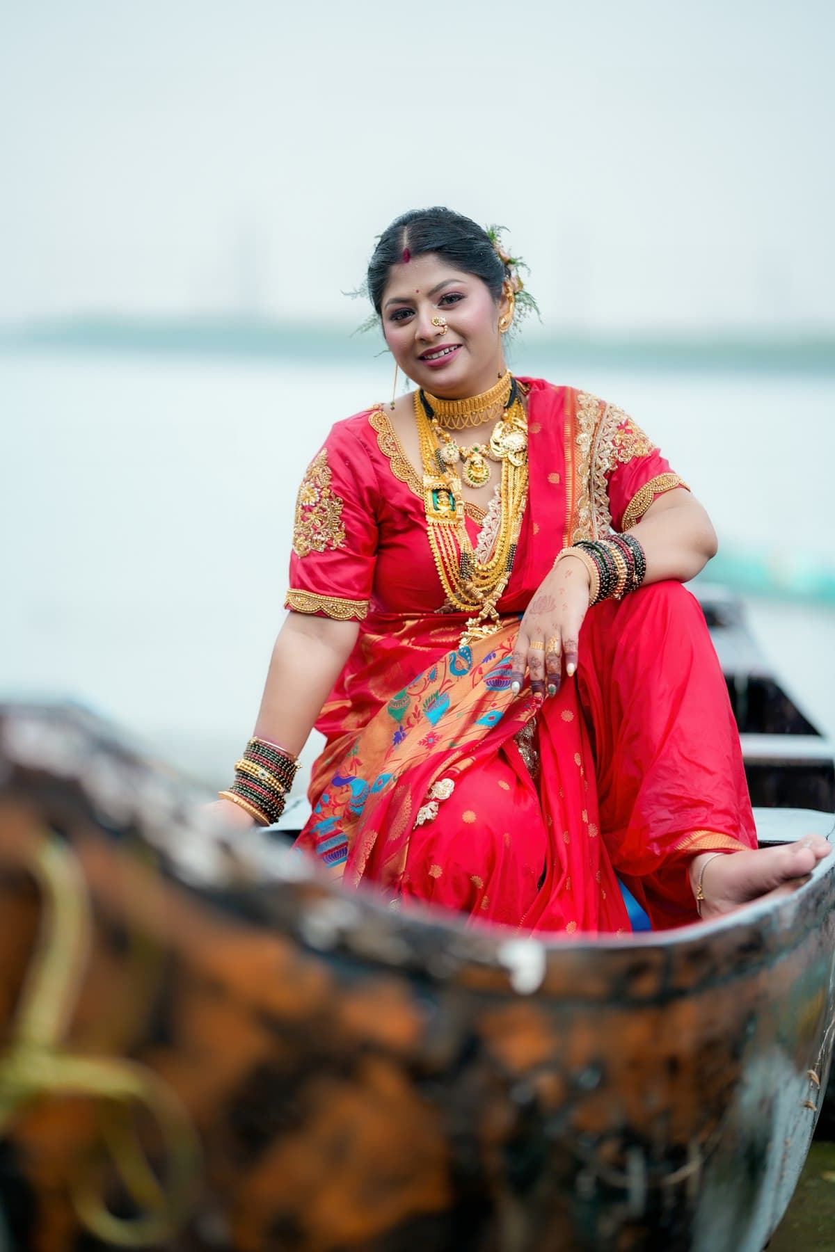 Smiling woman in a red silk saree and gold jewellery seated in a wooden boat