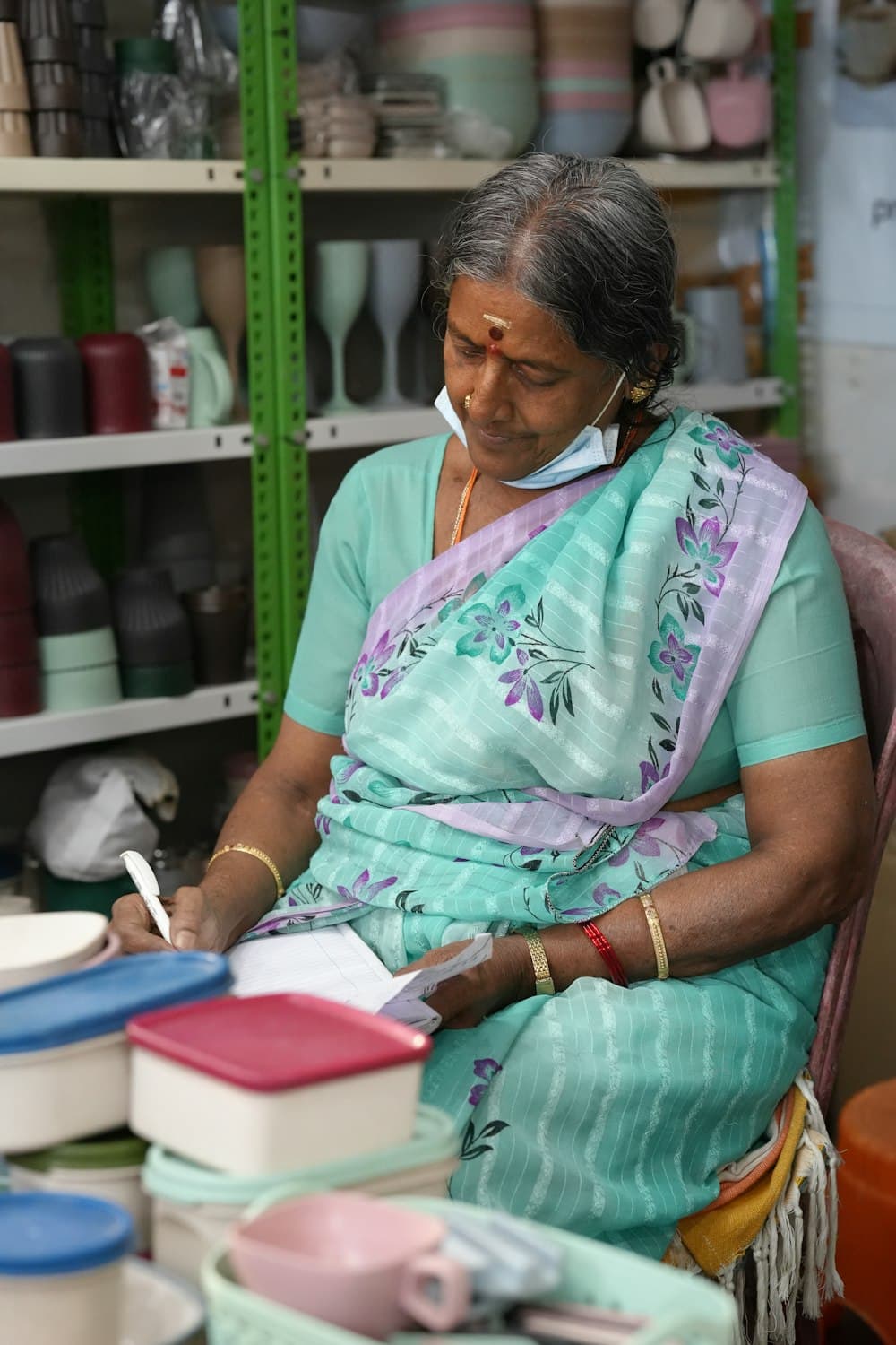 Woman in a pale blue saree writing in a notebook at her shop counter