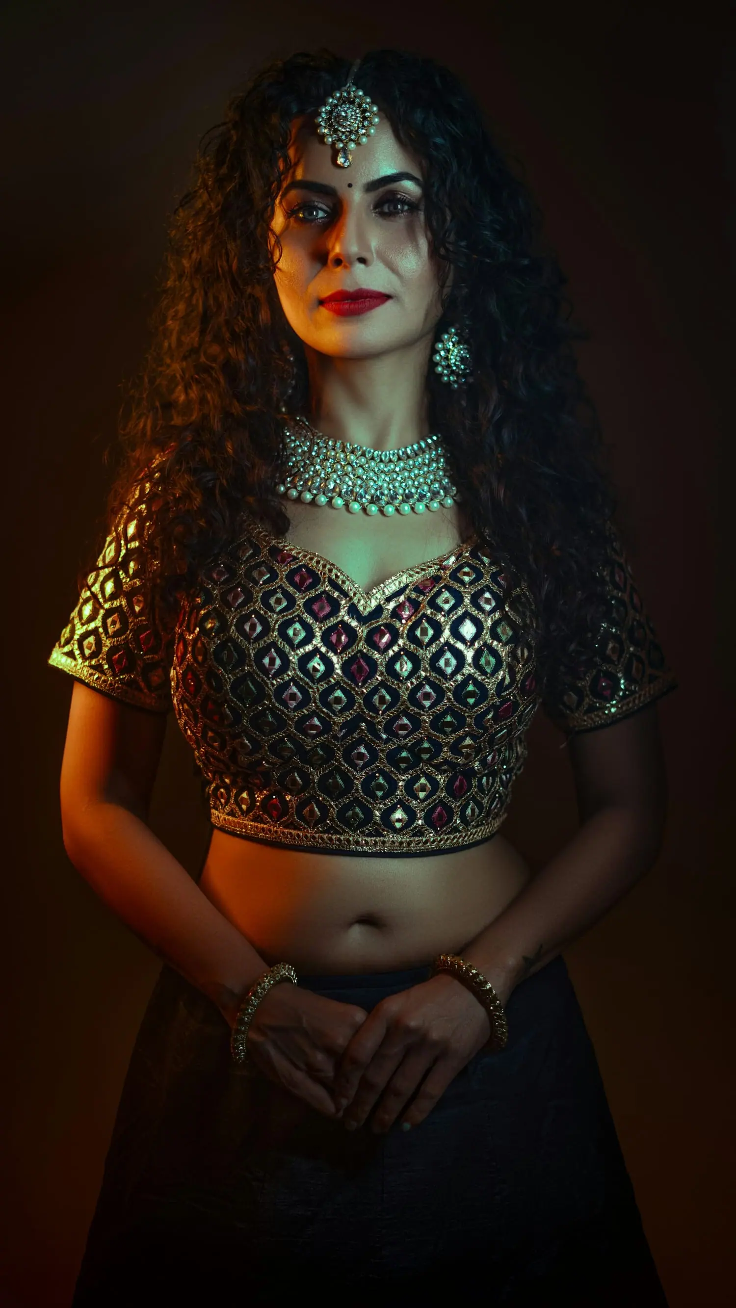 Woman with curly hair in a black and gold mirror-work choli and emerald jewellery, lit warmly against a dark backdrop